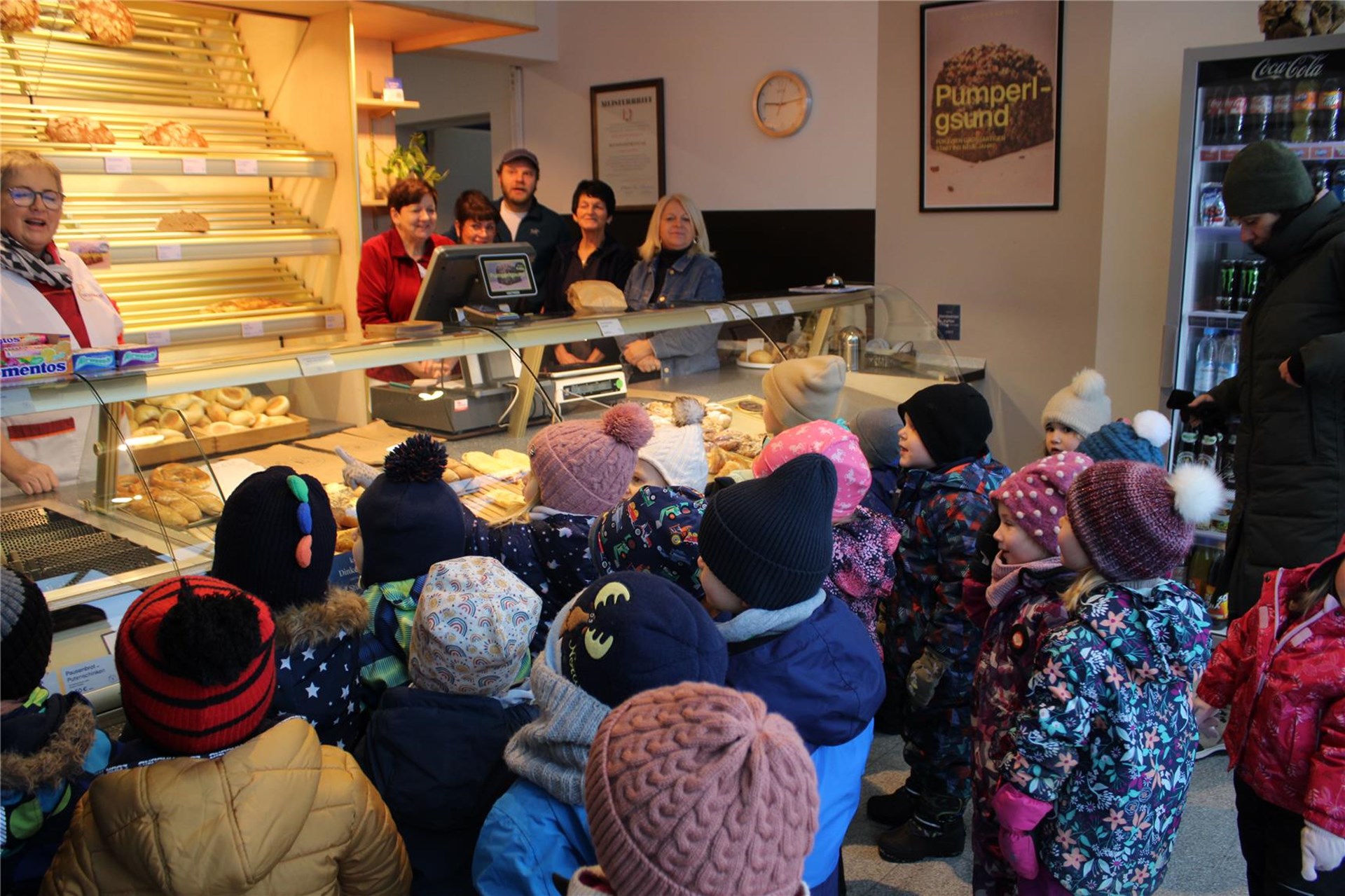 Eine Gruppe von Kindern in Winterkleidung besucht eine Bäckerei. Sie stehen vor der Theke und schauen auf die Backwaren.