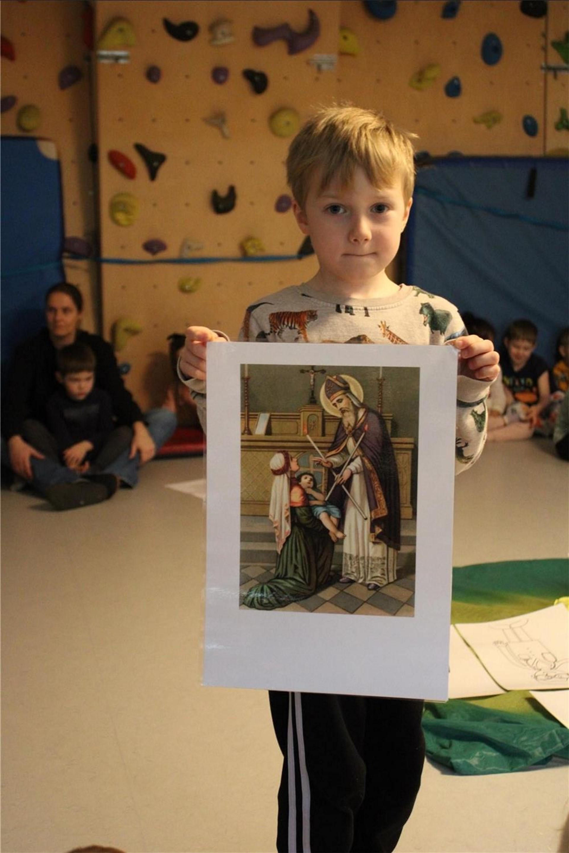 Ein Junge hält ein laminiertes Bild mit einer religiösen Szene hoch. Im Hintergrund sitzen andere Kinder und Erwachsene in einem Raum mit einer Kletterwand.