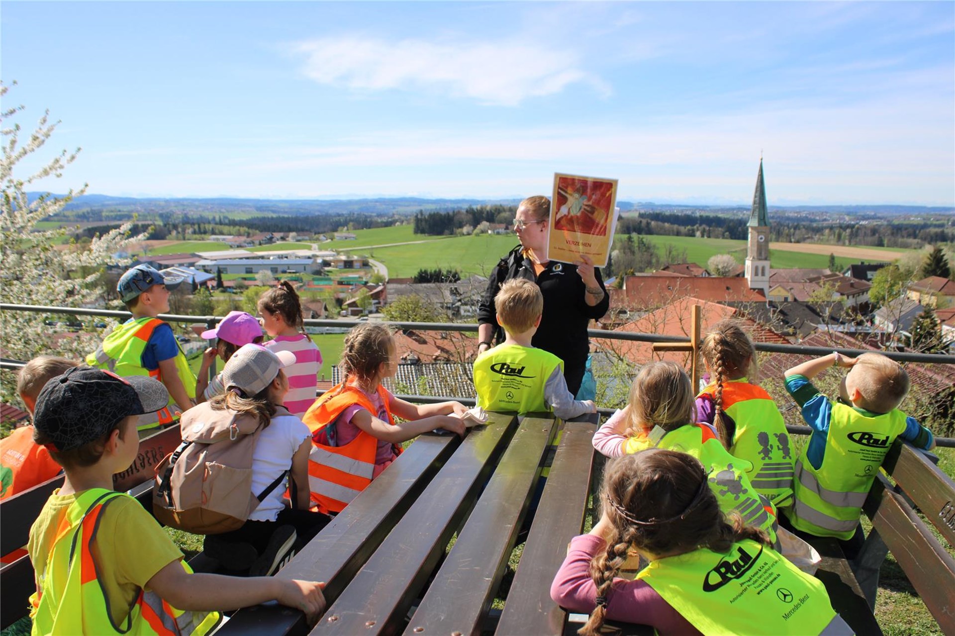 Eine Gruppe Kindergartenkinder in Warnwesten sitzt auf Bänken im Freien und lauscht einer Frau, die ein Bild mit der Aufschrift „Unsere Biene“ zeigt. Im Hintergrund erstreckt sich eine weite Landschaf