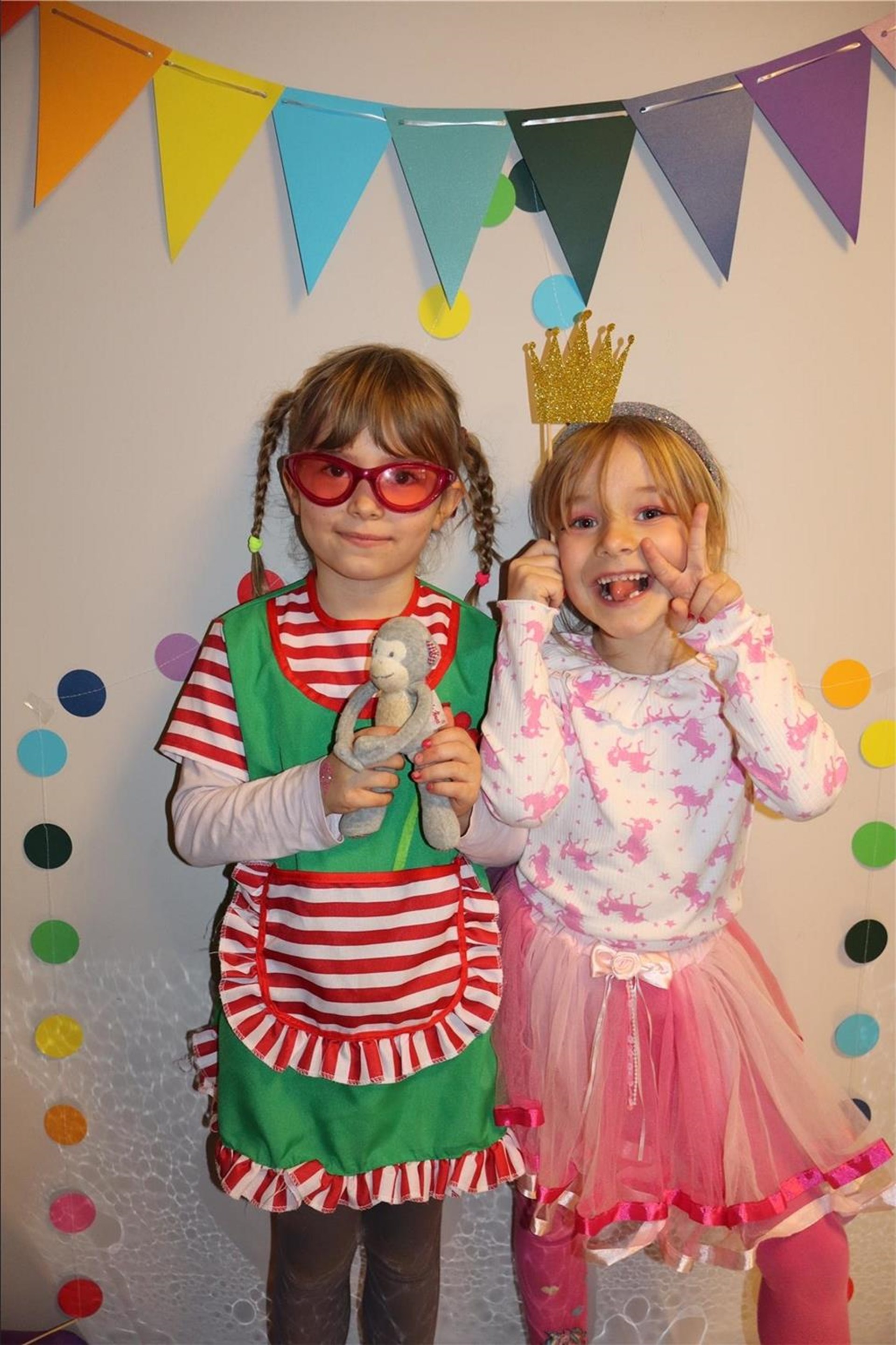 Zwei Kinder in bunten Faschingskostümen stehen vor einer Wand mit Wimpelgirlande. Eines trägt ein rot-grünes Kleid mit Plüschaffe, das andere ein pinkes Tutu, Krone und zeigt ein Peace-Zeichen.