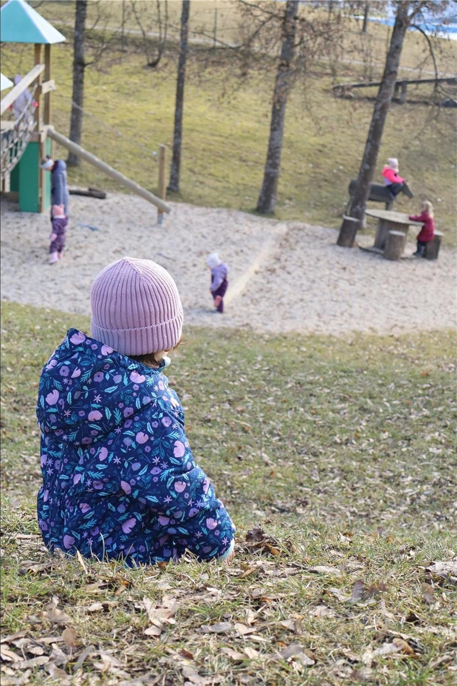 Ein Kind in einer blauen Winterjacke mit Blumenmuster und einer rosa Strickmütze sitzt auf einer Wiese und schaut zum Spielplatz. Im Hintergrund spielen mehrere Kinder im Sandbereich und an Tischen