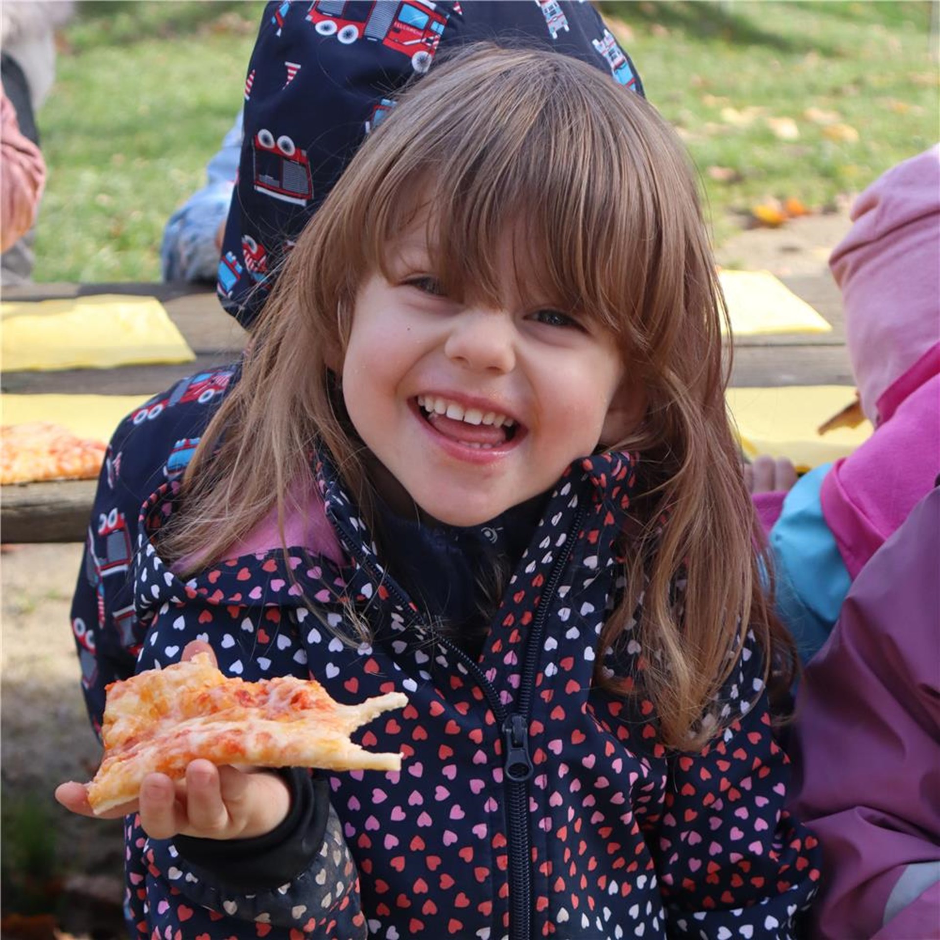Ein Mädchen hält ein Stück Pizza in der Hand und lächelt dabei.