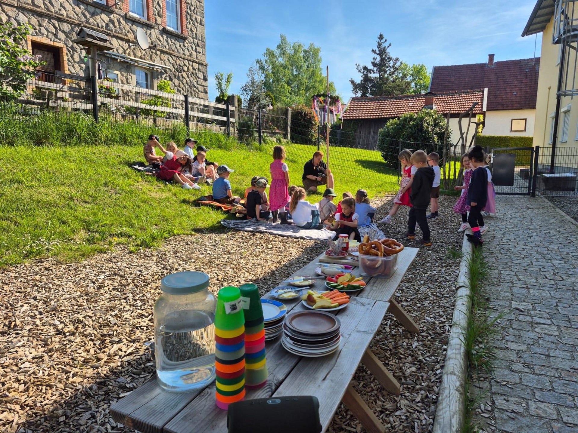 Kinder feiern im sonnigen Garten eines Kindergartens ein Maifest: Einige sitzen auf Picknickdecken im Gras, während andere spielen oder in Trachten herumlaufen. Im Vordergrund steht ein gedeckter Holztisch mit bunten Bechern, Tellern, Wasserkrug, Gemüsesticks, Dips, Obst und einer großen Schüssel mit Brezeln.